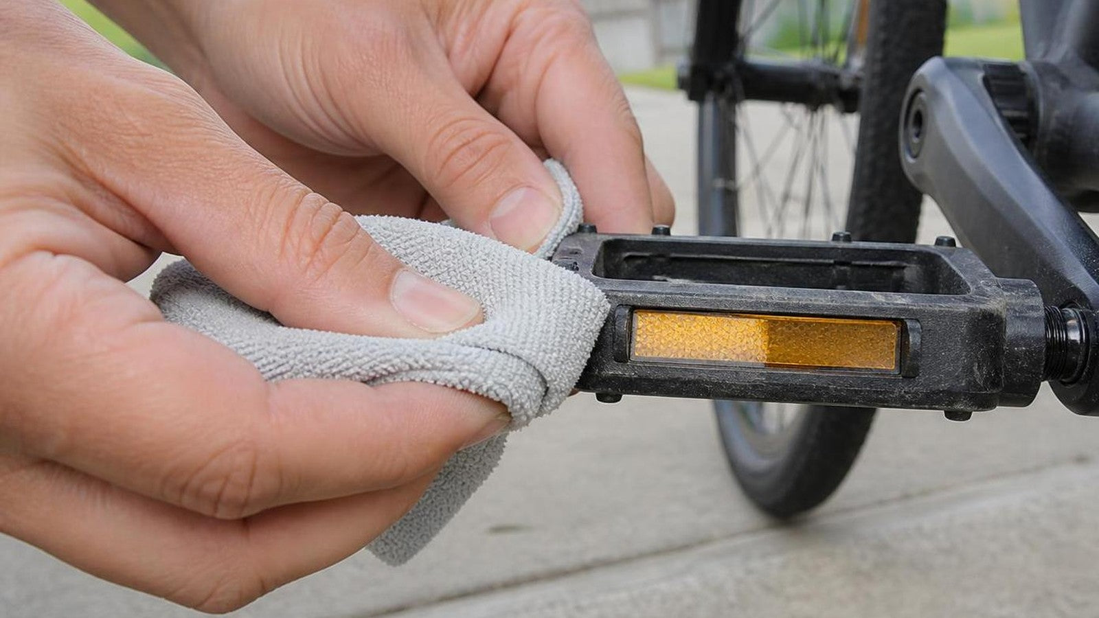 Bare hands wiping the amber reflector on a black flat pedal with a gray microfiber cloth on a driveway