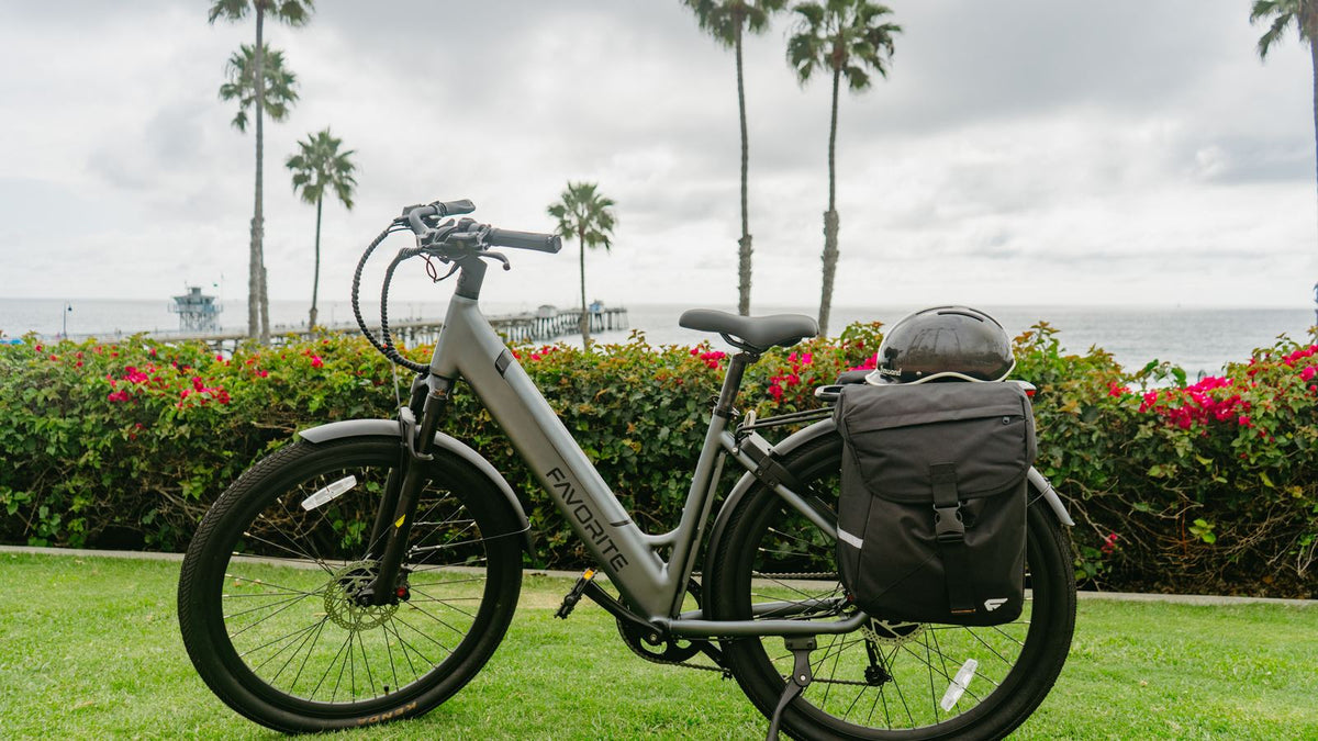 FavoriteBikes step-through electric bike parked on grass with a rear pannier storage bag and helmet, coastal path in the background