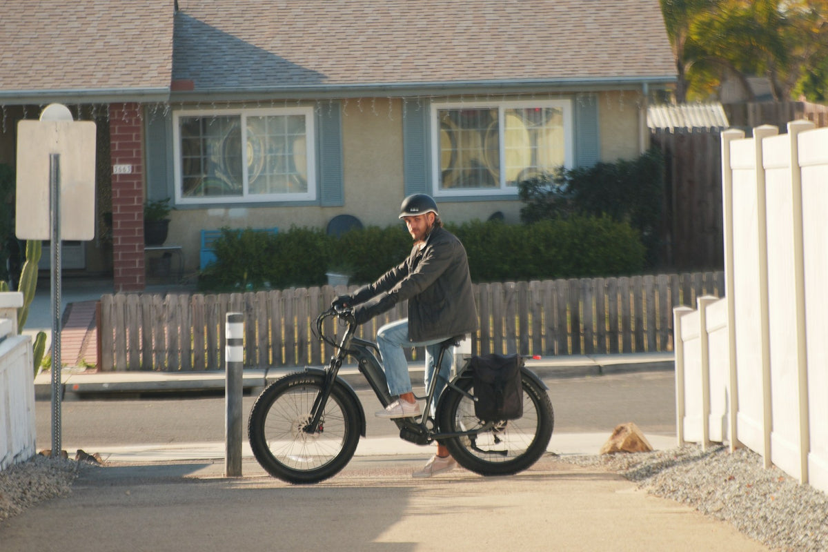 A helmeted person standing over a dark bicycle beside a wooden fence and a house, with a black bag at the rear.