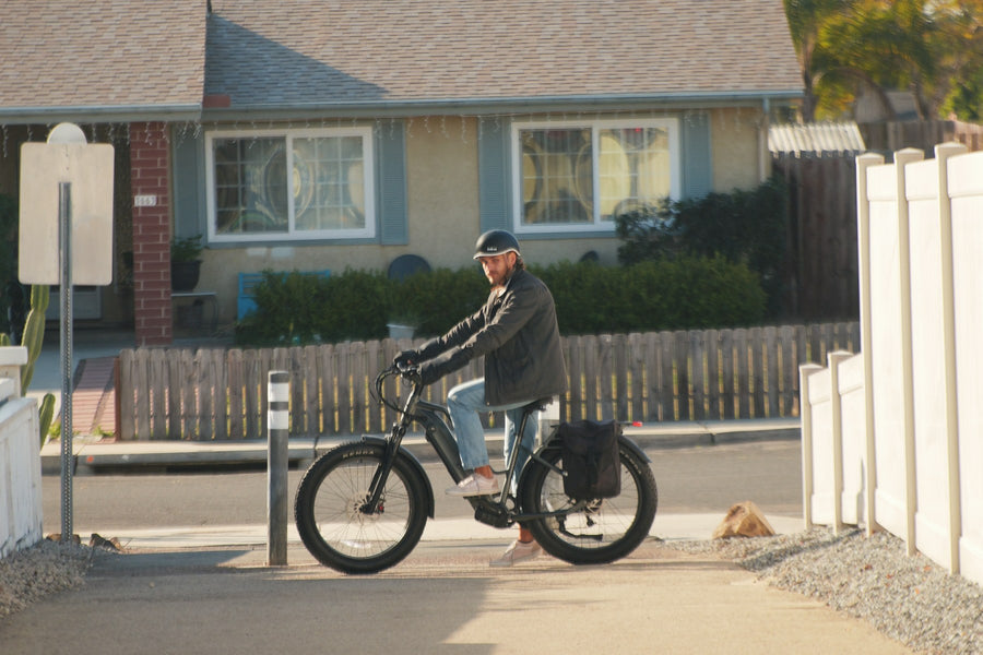 A helmeted person standing over a dark bicycle beside a wooden fence and a house, with a black bag at the rear.