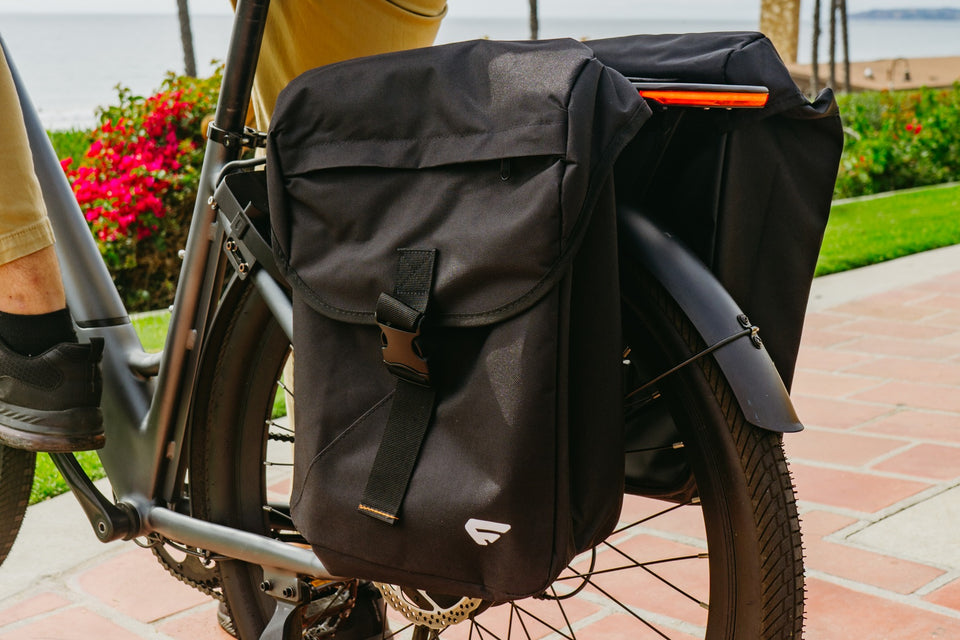 Close-up of black rear panniers beside a bicycle wheel on a brick path, with flowers and the coast in the background.