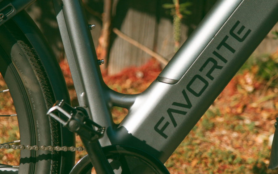 Close view of a gray e-bike frame with FAVORITE lettering, pedal and rear wheel against flowering plants.