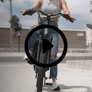 Front view of a person riding a dark grey electric bike costco from FavoriteBikes on a concrete surface. The rider is wearing a striped crop top, ripped blue jeans, and white sneakers. The costco e bike features a robust frame, wide tires, and a bright headlight, and the rider appears to be enjoying a casual ride in an urban setting