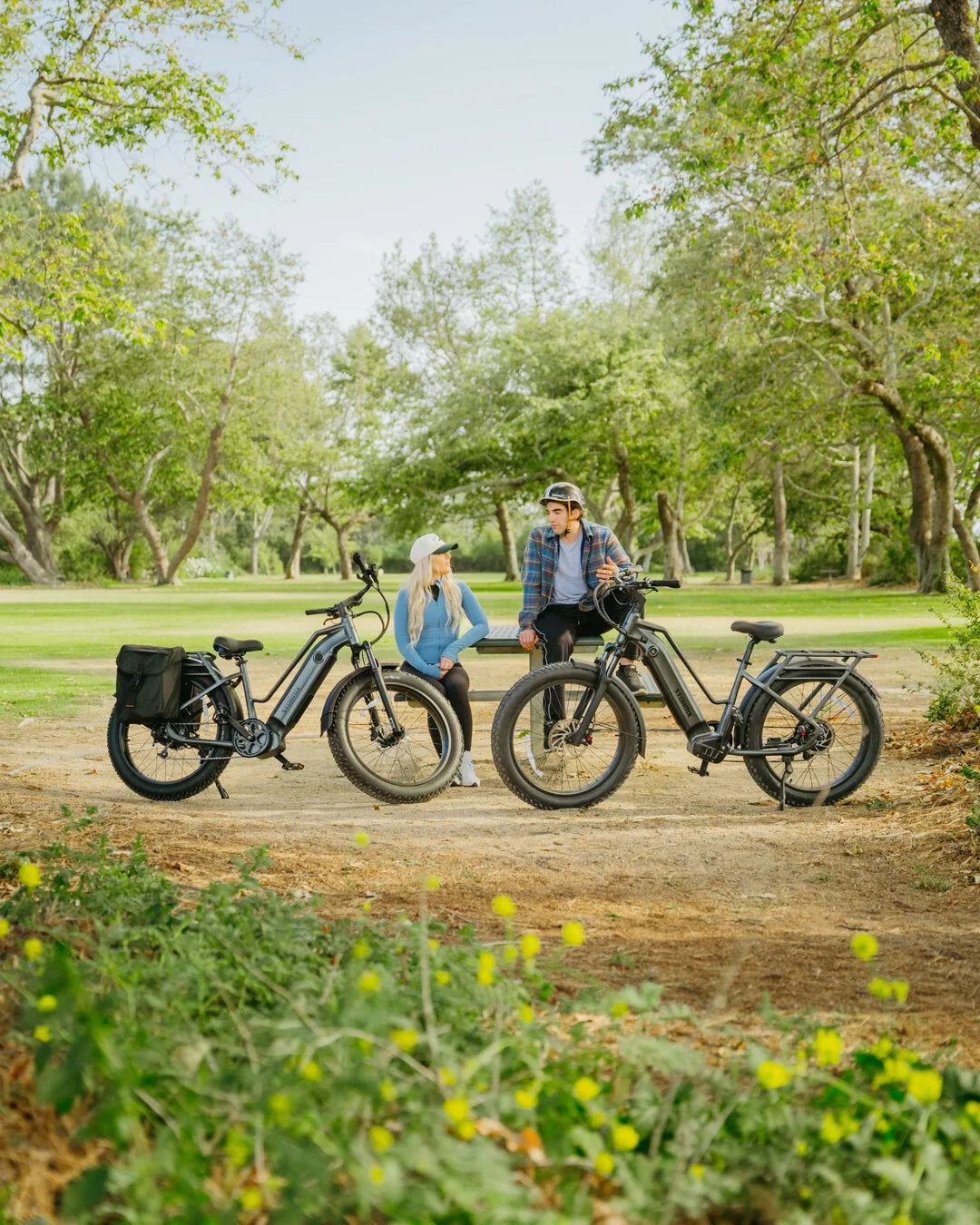 Two helmeted adults sit between two gray StormX fat tire e-bikes in a wooded park