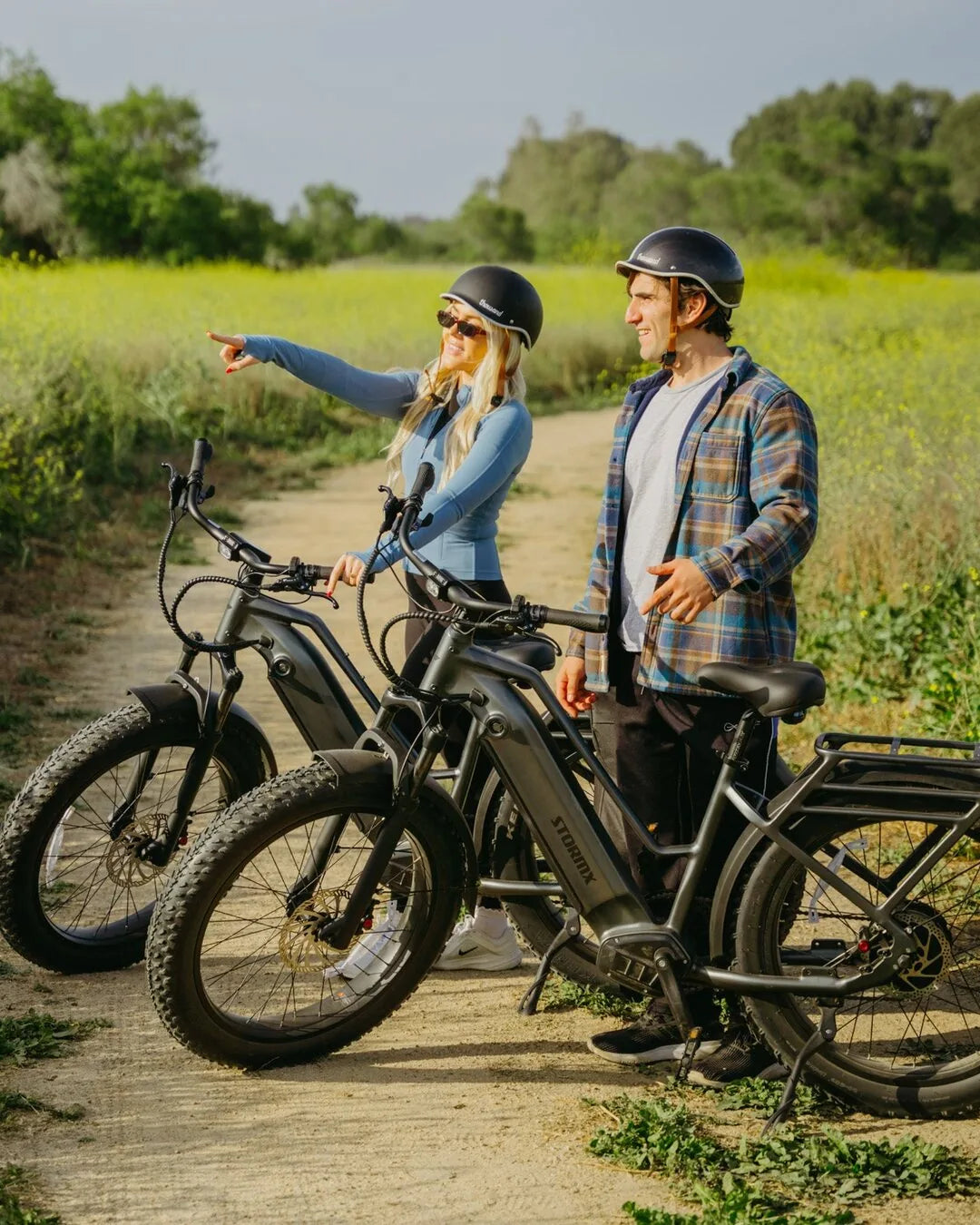 Two helmeted adults stand with gray StormX fat tire e-bikes on a dirt trail as one points